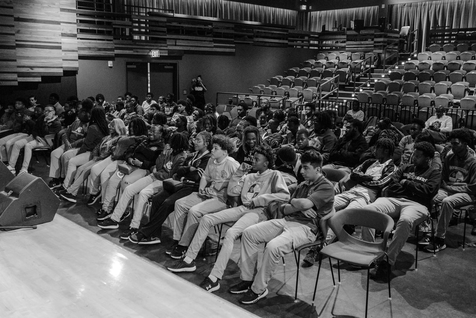 Students in school uniforms seated in a theater, listening to a performance